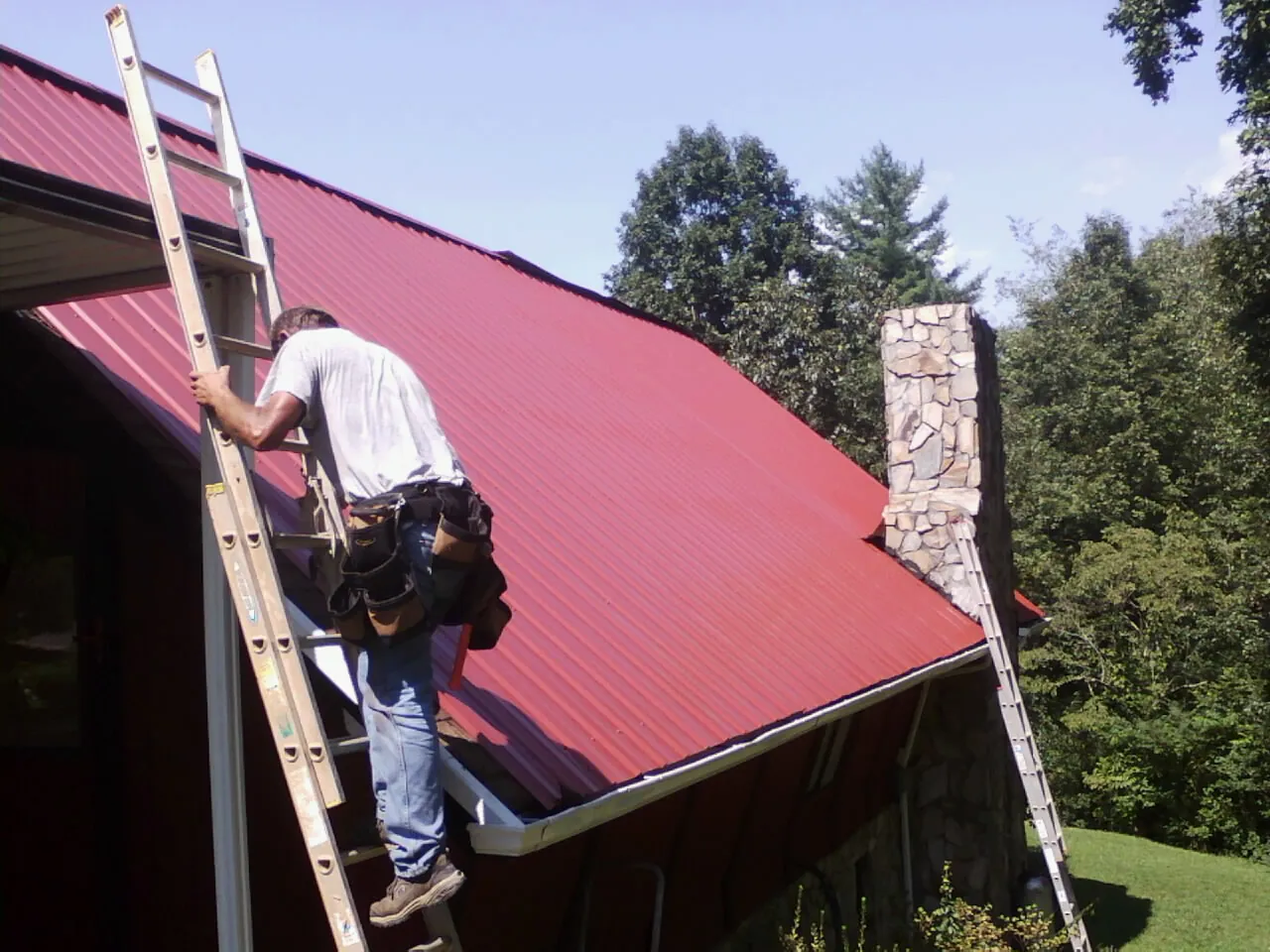 Roofer on a ladder beside a red metal roof and stone chimney — Soffit & Fascia Repair by Roofing Brothers in North Dighton